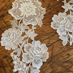 May include: White lace applique with a floral design. The applique features a large flower with smaller flowers and leaves surrounding it.