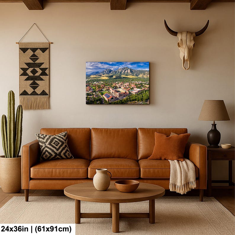 May include: A canvas print of a scenic landscape featuring a town nestled at the base of majestic mountains. The print, measuring 24x36 inches, showcases a vibrant scene with lush greenery, buildings, and a clear blue sky. The artwork is displayed above a brown leather sofa.