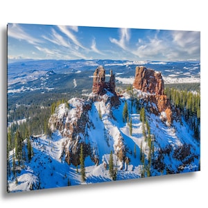 May include: A scenic landscape photograph featuring two large, reddish-brown rock formations covered in snow. The image captures a winter scene with evergreen trees and a bright blue sky with wispy clouds. The photograph is mounted on a black frame.