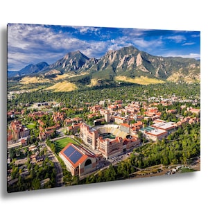 May include: An aerial view of a university campus nestled at the base of majestic mountains under a blue sky with scattered clouds. The campus features buildings with red-tiled roofs, a stadium, and lush green spaces, creating a picturesque landscape.