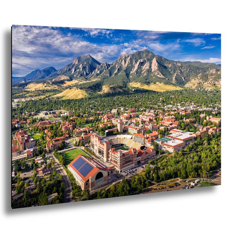 May include: An aerial view of a university campus nestled at the base of majestic mountains under a blue sky with scattered clouds. The campus features buildings with red-tiled roofs, a stadium, and lush green spaces, creating a picturesque landscape.