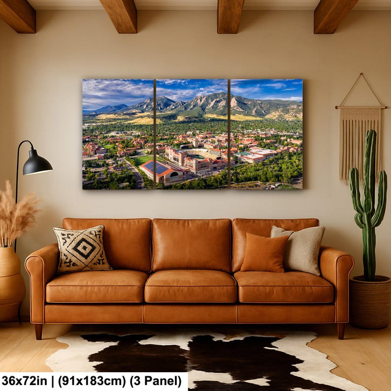 May include: A three-panel canvas print depicting a scenic aerial view of a city nestled at the base of majestic mountains under a blue sky. The artwork is 36x72 inches (91x183cm) and hangs above a brown leather sofa.