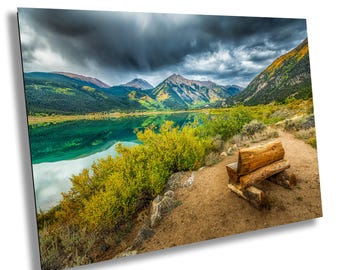 May include: A wooden bench sits on a path overlooking a lake surrounded by mountains. The sky is cloudy and the water is a vibrant green.