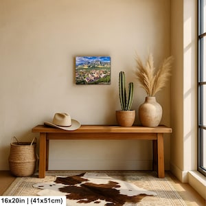 May include: A framed landscape print of a town nestled in a valley with mountains in the background. A tan hat rests on a wooden bench, with a woven basket and a cowhide rug in front. A cactus and vase with dried plants are also present.