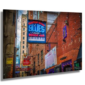 May include: A vibrant street scene featuring brick buildings adorned with neon signs. The prominent sign reads "Bourbon Street Blues & Boogie Bar" with a piano keyboard graphic. String lights and other signs add to the lively atmosphere, creating a classic bar and music venue vibe.