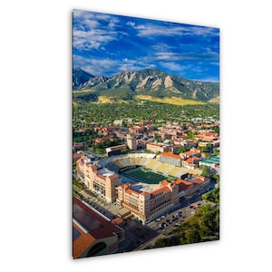 Peut inclure: Vue aérienne d'un campus universitaire avec un stade de football, entouré de bâtiments aux toits de tuiles rouges. Des montagnes et un ciel bleu nuageux en arrière-plan. Le stade a un terrain vert.