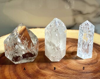 May include: Three clear quartz crystal points, labeled A, B, and C, on a wooden surface. The crystals have a unique, cloudy, and textured appearance.