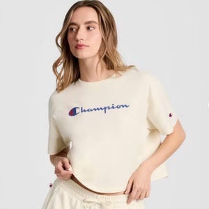 May include: Cream-colored Champion t-shirt with the brand name in blue script across the chest. A small Champion logo is on the left sleeve. The shirt is paired with matching cream-colored pants.