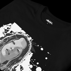 Könnte beinhalten: Schwarzes T-Shirt mit einem weißen Grafikdruck eines Frauen Gesichts mit Spritzlack-Akzenten. Das Etikett lautet "nlshirt."