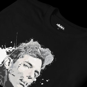 Könnte beinhalten: Schwarzes T-Shirt mit einem weißen und grauen Grafikdruck eines Männergesichts mit einem Farbspritzer-Effekt. Der Text "n1tshirt." befindet sich auf dem Etikett.