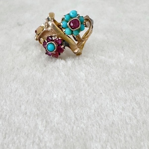 May include: A gold-toned ring featuring two floral designs. One flower has turquoise stones, the other has ruby stones, each with a central stone. The ring is set on a white surface.