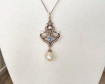 Könnte beinhalten: Eine goldfarbene Anhängerkette mit einem verzierten Design. Der Anhänger verfügt über kleine Perlen, einen blauen Edelstein und eine große tropfenförmige Perle. Die Halskette hängt an einer filigranen Kette.