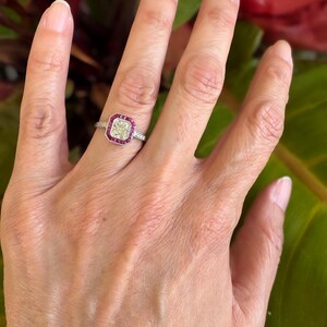 May include: A silver-toned ring featuring a rectangular center stone with a pale yellow hue, surrounded by a halo of small, vibrant red stones. The band is thin and metallic, with small, clear stones embedded along the sides of the ring shank.
