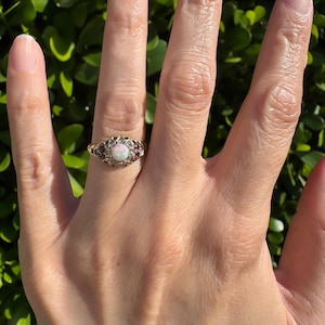 May include: A gold ring with an oval, iridescent opal center stone, surrounded by small red gemstones. The ring has an ornate, filigree design on the band. The ring is shown on a hand.