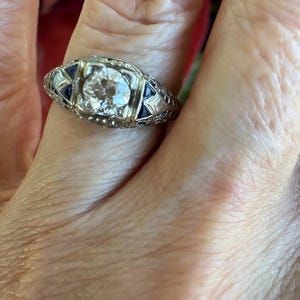 May include: A vintage-style ring with a large, round, clear gemstone at the center. The ring has a silver-colored band with intricate detailing and small, dark blue accent stones on either side of the center stone.