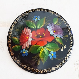 May include: A round black brooch featuring a hand-painted floral arrangement with a large central red flower, pink and purple blossoms, and small blue flowers surrounded by green leaves. The edge has a gold-coloured decorative scroll pattern.