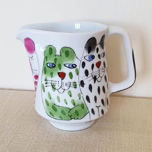 Può includere: Una lattiera in ceramica bianca con illustrazioni dipinte a mano di tre gatti stilizzati. Il gatto centrale è verde con occhi blu, affiancato da un gatto rosa a sinistra e un gatto bianco con macchie nere sulla destra. Ha un manico.