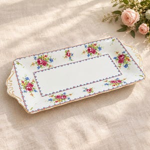 Peut inclure: Un plateau de service rectangulaire en céramique blanche orné d'un motif floral style point de croix avec des roses roses et des fleurs bleues. Le plateau présente une bordure décorative, des anses festonnées dorées.