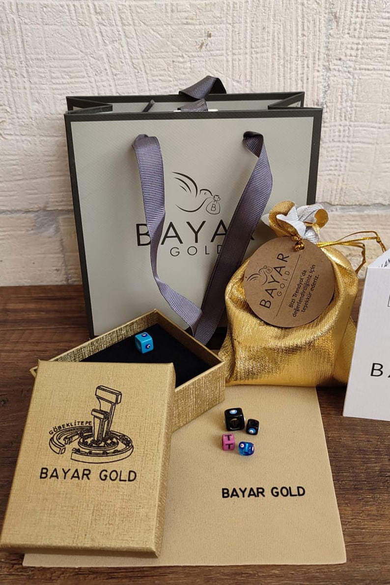 May include: A gold gift bag with a white ribbon and a brown tag that says "BAYAR GOLD" and "Bizi Trendyol'da değerlendirdiğiniz i&ccedil;in teşekk&uuml;r ederiz."  The bag is sitting on a brown surface with a gold box and a small gold pouch with a brown tag that says "BAYAR GOLD".