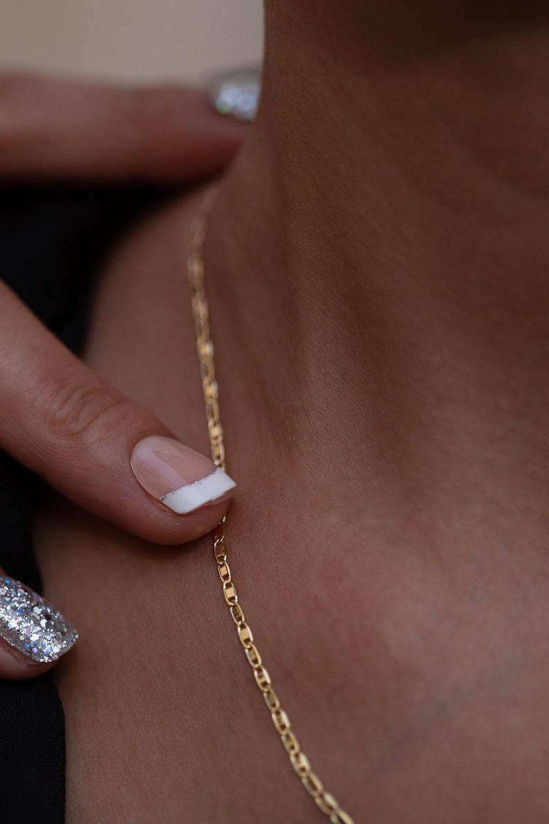 May include: A close-up of a gold chain necklace worn by a person with a white manicure. The chain is a delicate, thin, and simple design.