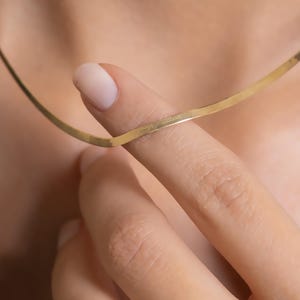 Puede incluir: Un delicado collar de color dorado con un diseño plano y delgado. El collar está sostenido por un dedo, mostrando su sutil elegancia. La joya parece estar hecha de un metal brillante, que refleja la luz.