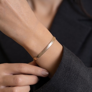 Puede incluir: Un brazalete dorado con un patrón de espiga. El brazalete se lleva en la muñeca, y la imagen es un primer plano. El brazalete tiene un acabado liso y pulido y un diseño clásico. El fondo es de color oscuro y sólido.