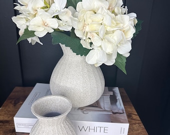 Può includere: Due vasi in ceramica grigia con una superficie testurizzata. Il vaso più grande contiene un mazzo di ortensie bianche. I vasi sono appoggiati su un libro intitolato "White" con il testo "The White Company" visibile sul dorso.
