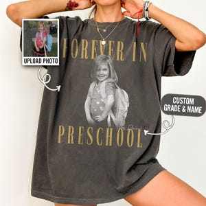 Może przedstawiać: Ciemnoszary t-shirt oversize z czarno-białym zdjęciem dziecka z plecakiem. Napis "FOREVER IN" znajduje się nad zdjęciem, a "PRESCHOOL" poniżej, wykonany złotymi literami. Małe wstawione zdjęcie przedstawia to samo dziecko.