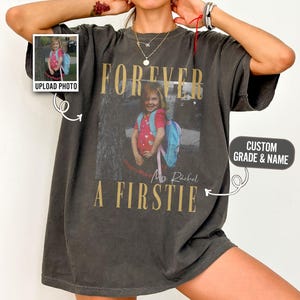 Może przedstawiać: Ciemnoszary t-shirt oversize z grafiką w stylu vintage przedstawiającą dziecko z plecakiem. Napis głosi FOREVER A FIRSTIE oraz Mrs. Rachel. Projekt zawiera małe okienko na zdjęcie z napisem UPLOAD PHOTO oraz pole tekstowe z napisem CUSTOM GRADE & NAME.