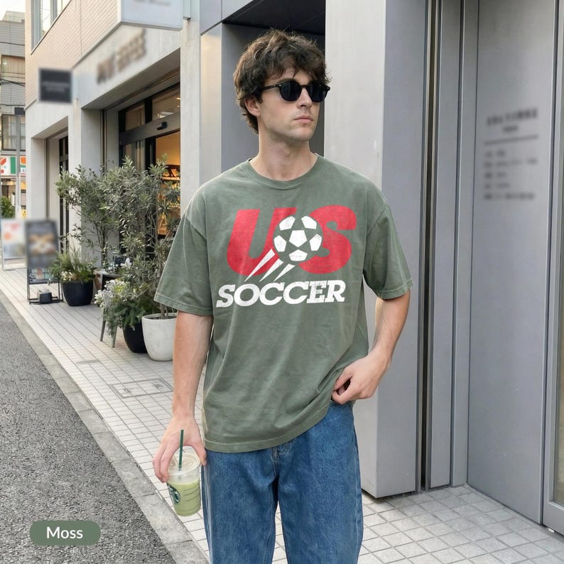 May include: A moss green short-sleeve t-shirt featuring a red graphic of the letters US with a white football in the centre and the word SOCCER printed in white underneath. The shirt is paired with blue denim jeans and black sunglasses.