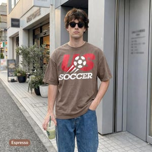 May include: A brown short-sleeve t-shirt featuring a graphic of a football and the red text US above the white text SOCCER. The shirt is paired with blue denim jeans. The design includes motion lines trailing behind the football graphic.