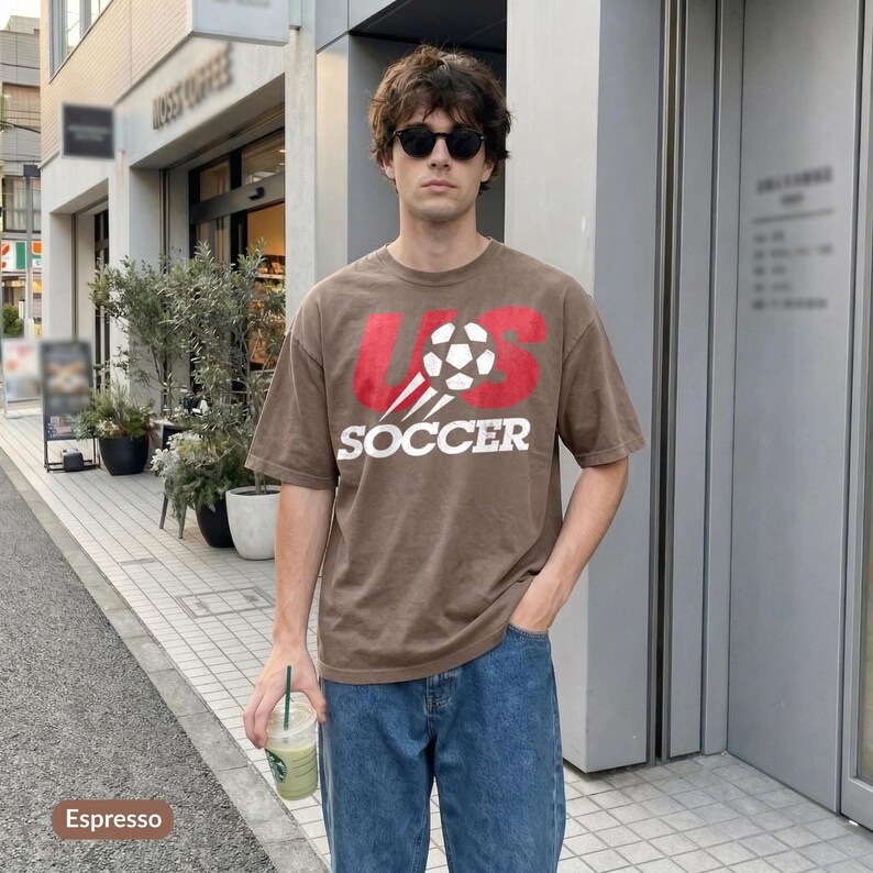 May include: A brown short-sleeve t-shirt featuring a graphic of a football and the red text US above the white text SOCCER. The shirt is paired with blue denim jeans. The design includes motion lines trailing behind the football graphic.