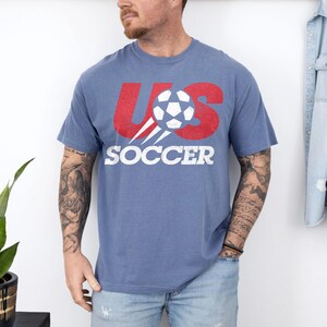 May include: A blue short-sleeved t-shirt featuring a distressed red graphic that reads US with a white football replacing the letter S. Below the graphic, the word SOCCER is printed in bold white capital letters across the chest area.