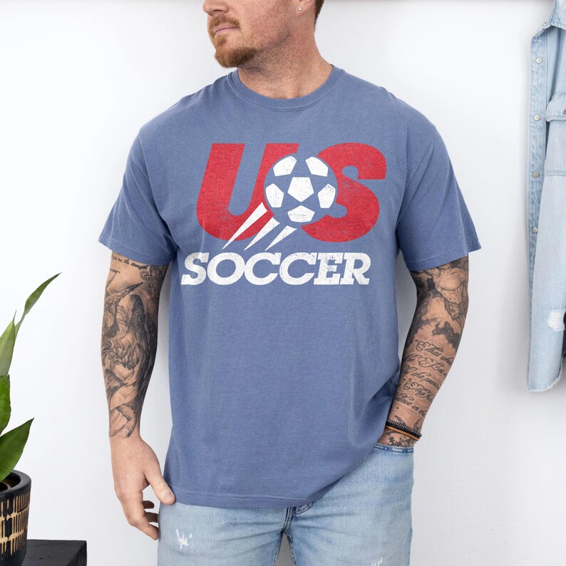 May include: A blue short-sleeved t-shirt featuring a distressed red graphic that reads US with a white football replacing the letter S. Below the graphic, the word SOCCER is printed in bold white capital letters across the chest area.