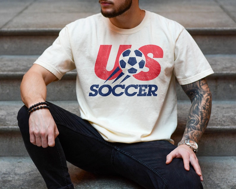 May include: A cream-coloured short-sleeve t-shirt featuring a graphic print on the chest. The design includes the red text US with a blue and white football replacing the letter S, and the word SOCCER in blue text underneath the graphic.