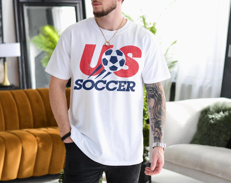 May include: A white short-sleeved t-shirt featuring a graphic on the chest with the red text US and the blue text SOCCER. A stylised football with blue and white panels is positioned between the letters, showing a distressed texture.