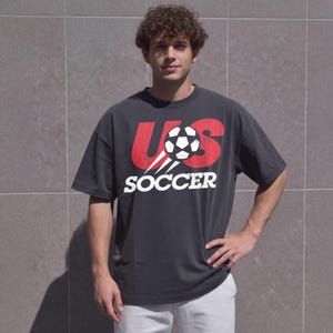 May include: A charcoal grey short-sleeve t-shirt featuring a large red graphic with the letters US and a black and white football in the centre. Below the graphic, the word SOCCER is printed in bold white capital letters.