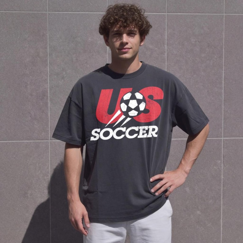 May include: A charcoal grey short-sleeve t-shirt featuring a large red graphic with the letters US and a black and white football in the centre. Below the graphic, the word SOCCER is printed in bold white capital letters.