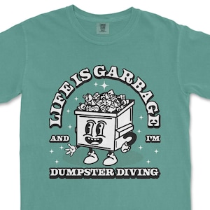 Peut inclure: T-shirt de couleur sarcelle avec un dessin rétro représentant un personnage de poubelle de dessin animé. Le texte indique "LIFE IS GARBAGE AND I'M DUMPSTER DIVING". La poubelle est remplie de déchets et a un visage souriant.