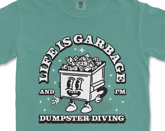 May include: Teal-colored t-shirt featuring a cartoon dumpster character with the text "LIFE IS GARBAGE AND I'M DUMPSTER DIVING" in a retro design. The dumpster is filled with trash and has a smiling face.
