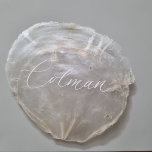 May include: A white seashell with the name "Colman" written in white calligraphy.