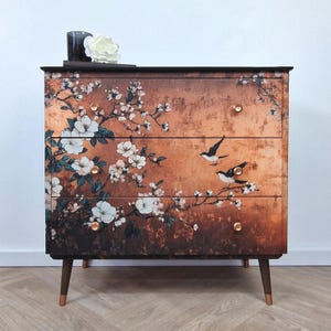 May include: A three-drawer chest of drawers with a floral and bird design. The chest has a dark brown top and legs with copper-coloured tips. The drawers feature white flowers and black birds against a copper-coloured background.