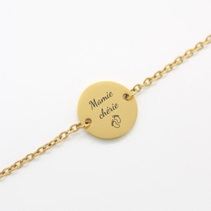 Puede incluir: Pulsera dorada con un colgante circular. El colgante tiene las palabras "Mamie chérie" y un diseño de huellas de pies. La pulsera está sobre un fondo blanco.