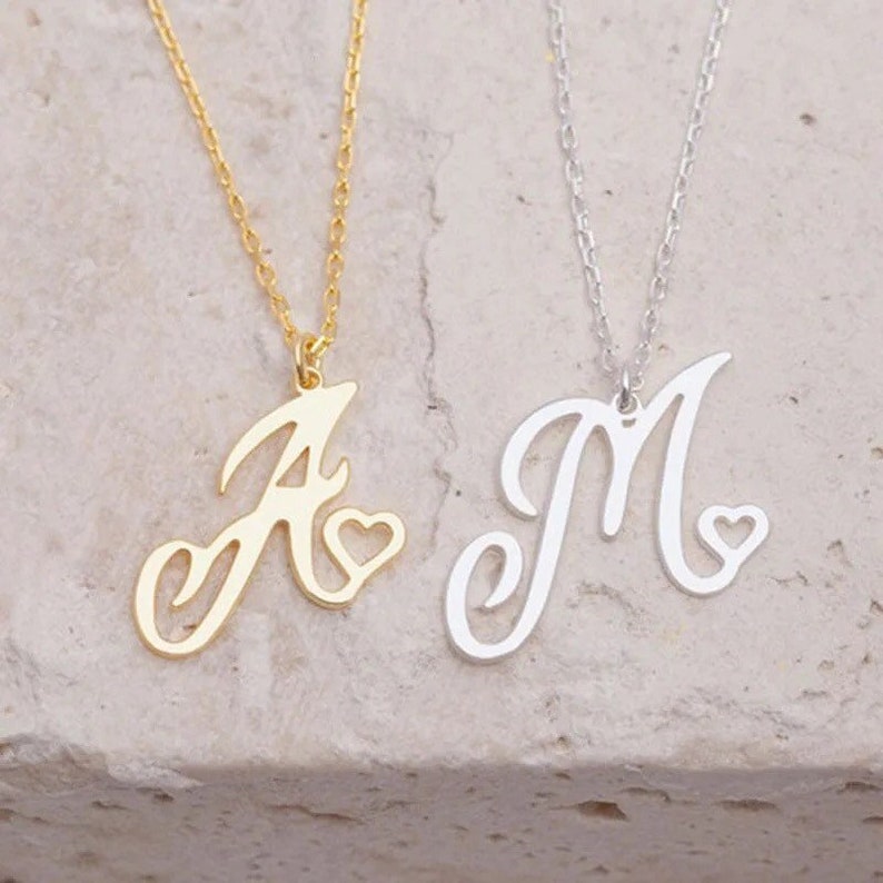 May include: Two gold necklaces with cursive letter pendants and small heart charms. The necklace on the left has the letter "A" and the necklace on the right has the letter "M".
