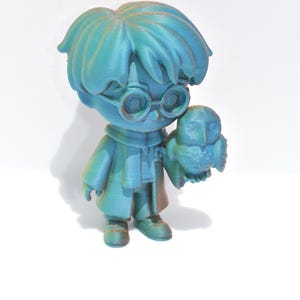 May include: A teal-colored figurine of a character with round glasses, holding a small owl. The figure is wearing a coat and has a detailed hairstyle. The figurine is approximately 4 inches tall.