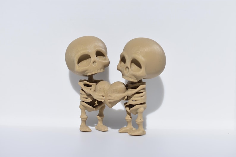 May include: Two small, beige skeleton figurines holding a heart-shaped object. The skeletons are posed as if they are embracing.