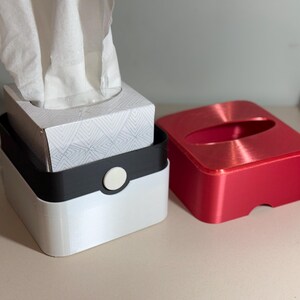 May include: A set of tissue boxes in white, black, and red. The white box has a black band with a white button. The red box has a rounded oval opening. White tissues are visible.