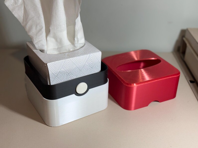May include: A set of tissue boxes in white, black, and red. The white box has a black band with a white button. The red box has a rounded oval opening. White tissues are visible.