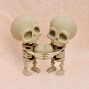 May include: Two small skeleton figurines holding a heart-shaped object. The skeletons are made of a light brown material and have a detailed skeletal structure. The heart is also made of a light brown material and has a textured surface.