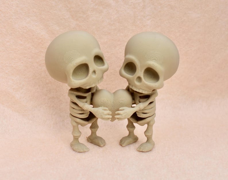 May include: Two small skeleton figurines holding a heart-shaped object. The skeletons are made of a light brown material and have a detailed skeletal structure. The heart is also made of a light brown material and has a textured surface.